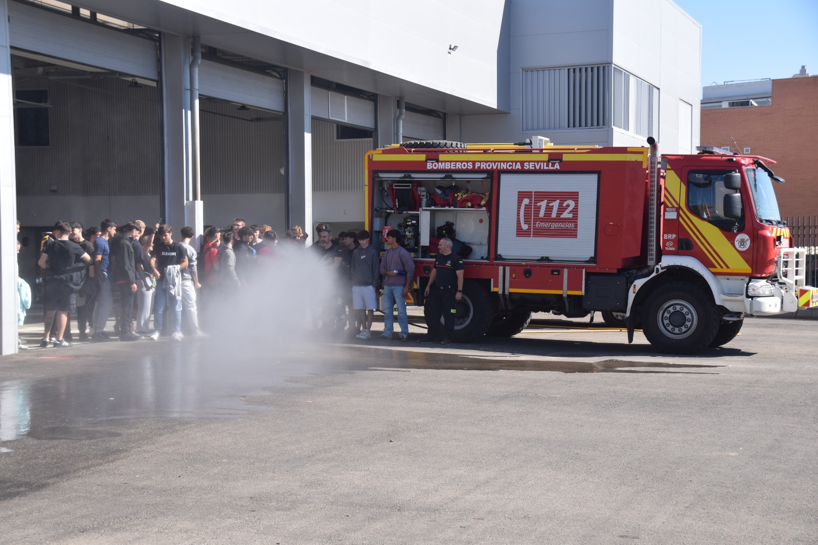 Un grupo de jóvenes junto a un camión de bomberos rojo con el rótulo 112 y una nube de vapor de agua en el suelo.