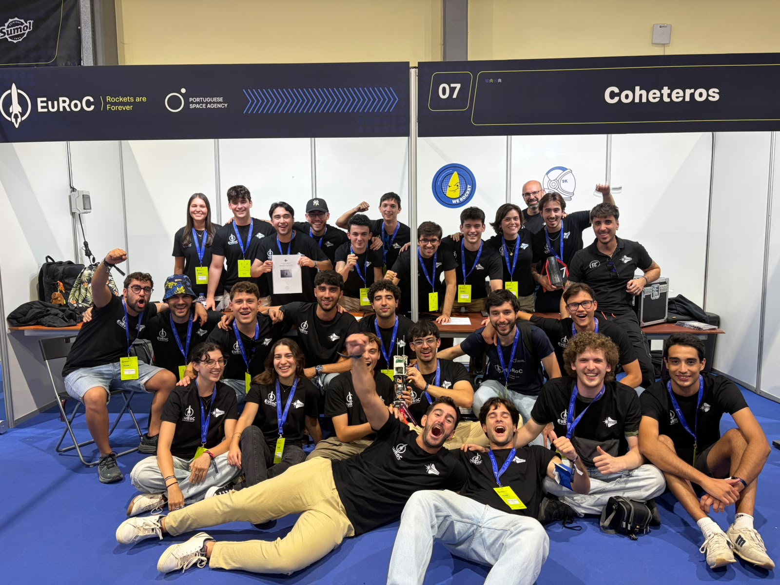 Un grupo numeroso de jóvenes con camisetas negras y acreditaciones azules posa sonriente en un stand de feria, algunos sentados en el suelo.