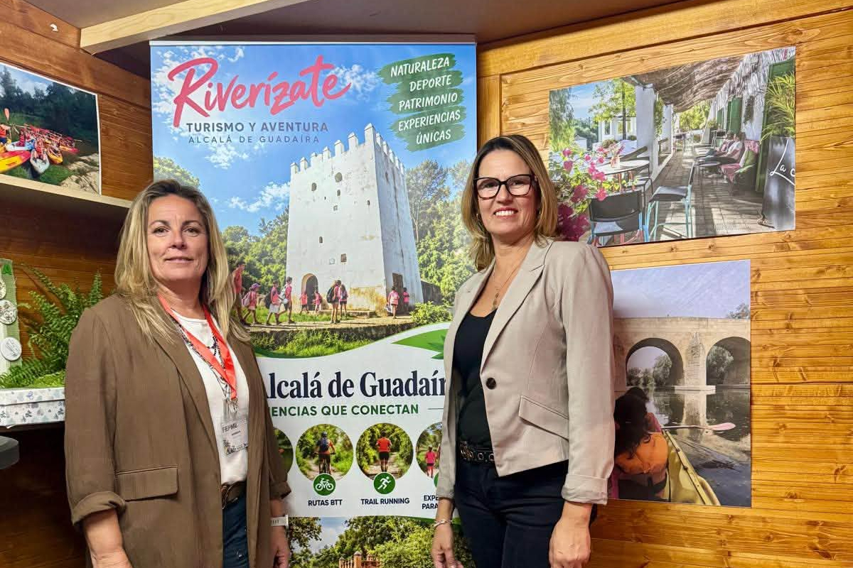 Dos mujeres de pie junto a un cartel de turismo y aventura de Alcalá de Guadaíra, en un stand de madera con fotografías.