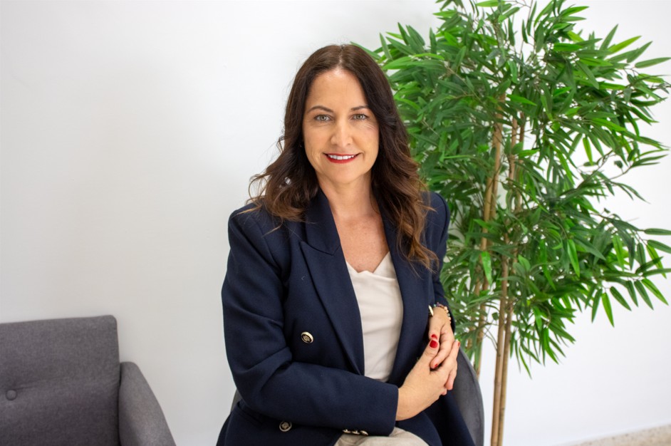 Mujer sonriente de pelo largo castaño, con chaqueta azul marino y blusa blanca, sentada junto a una planta y un sillón gris.