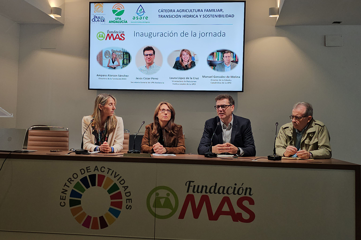 Cuatro personas sentadas tras una mesa con micrófonos, ante una pantalla que muestra el cartel de una jornada sobre agricultura y agua.