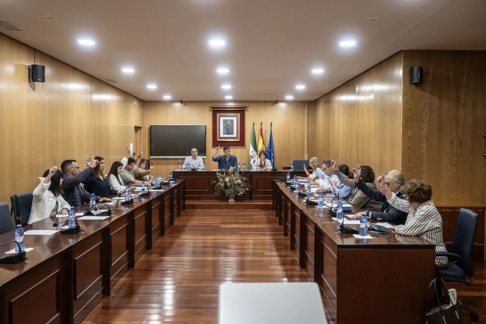 Varias personas sentadas en mesas largas de una sala de reuniones levantan la mano mientras tres presiden al fondo junto a banderas.
