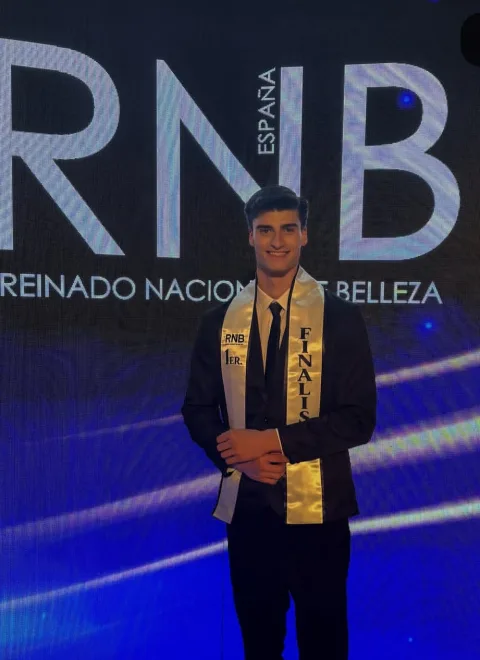 Un joven con traje y banda de finalista posa sonriente ante un fondo azul con las letras RNB España.