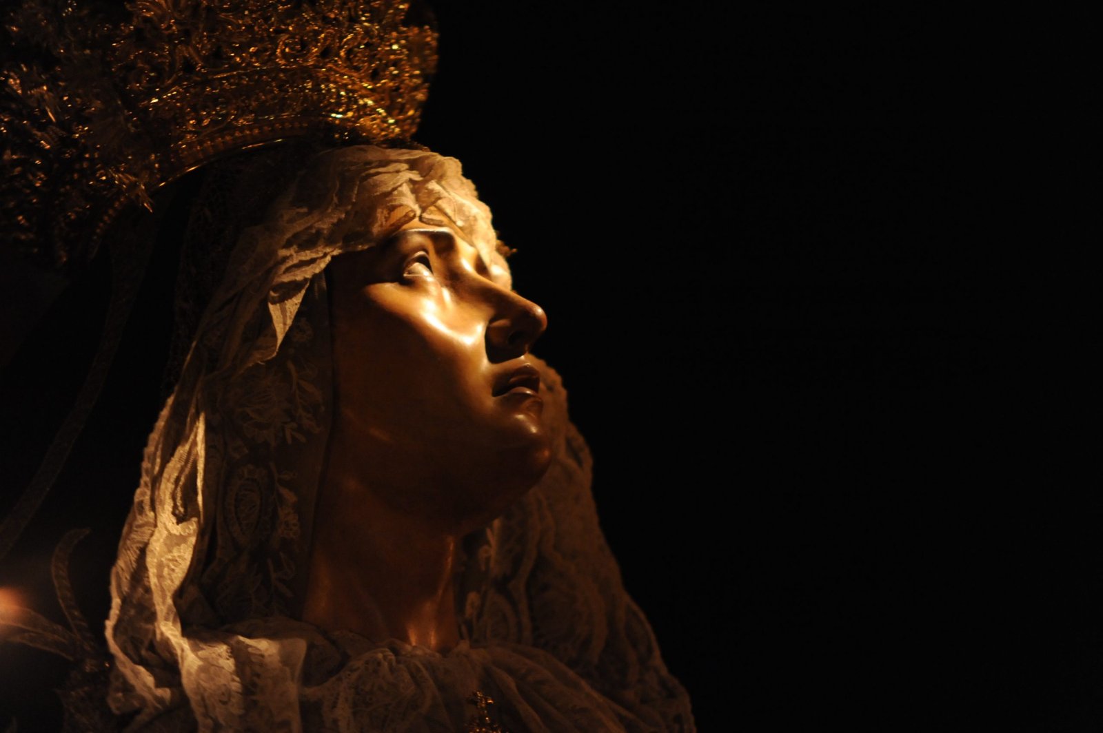 Rostro de una imagen religiosa femenina de perfil, con corona dorada y velo bordado, iluminado sobre fondo negro.