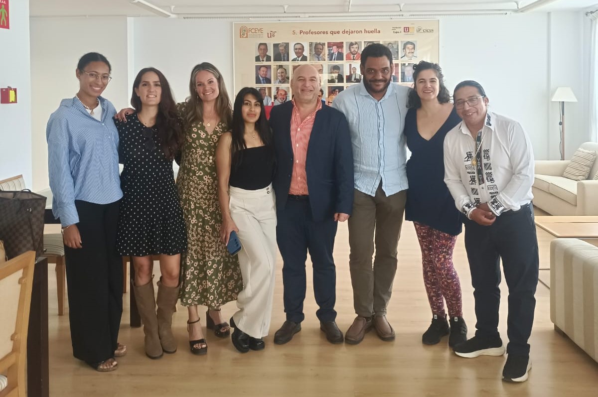 Ocho personas de pie, sonriendo en grupo, en una sala con un panel de fotos al fondo.