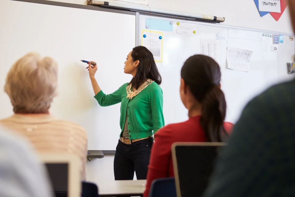 Una profesora con chaqueta verde escribe en una pizarra blanca ante varios alumnos sentados de espaldas en un aula.