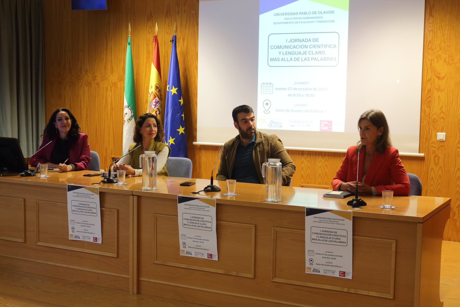 Cuatro personas sentadas tras una mesa en una sala con banderas y una pantalla de proyección al fondo.