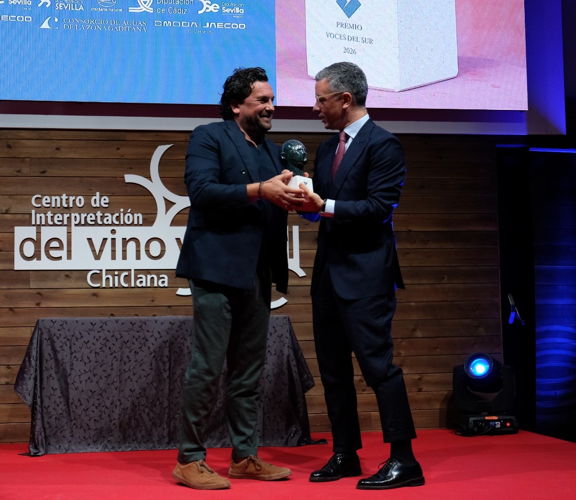 Dos hombres trajeados se dan la mano y sostienen un trofeo sobre una alfombra roja ante un panel de madera.