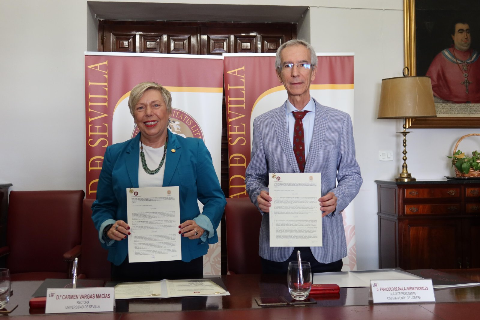 Una mujer y un hombre de traje sostienen sendos documentos delante de dos carteles de la Universidad de Sevilla, junto a una mesa con cartelas.