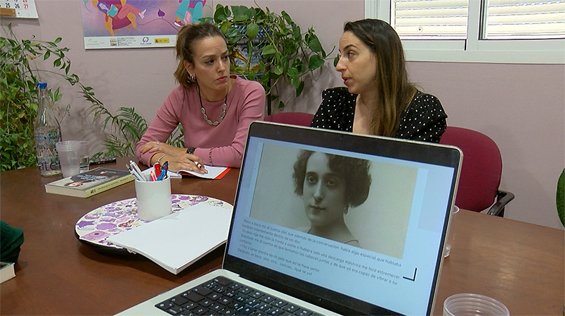 Dos mujeres sentadas a una mesa con un portátil que muestra el retrato de una mujer, junto a cuadernos y plantas.