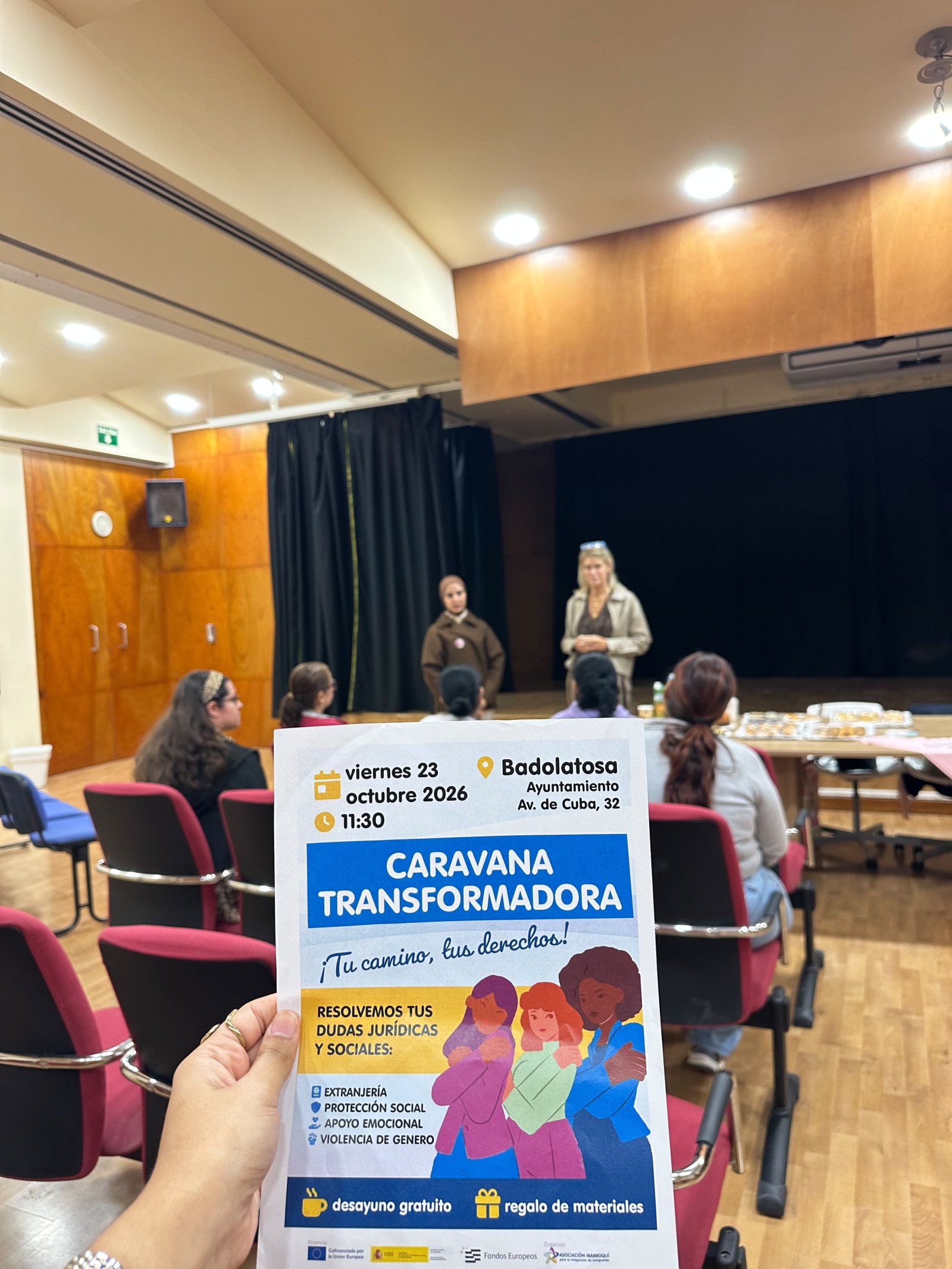 Una mano sostiene un folleto sobre una charla en un salón con sillas rojas y dos personas al fondo junto a una mesa con comida.