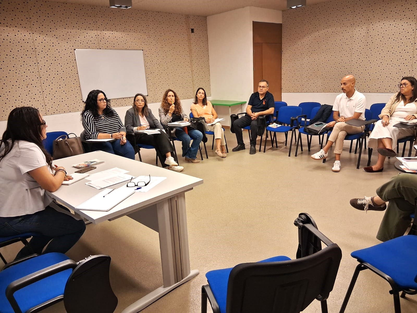 Un grupo de personas sentadas en sillas azules formando un círculo en una sala con mesa y pizarra blanca.