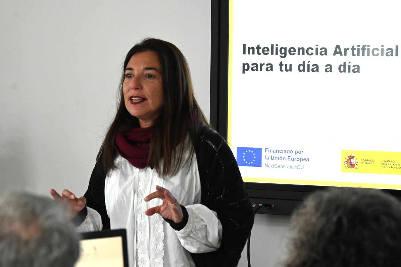 Una mujer habla gesticulando ante un público, junto a una pantalla que muestra el título Inteligencia Artificial para tu día a día.