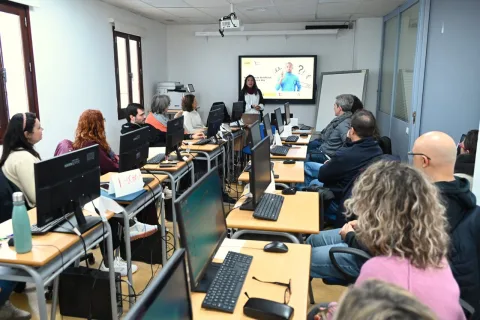 Personas adultas sentadas ante ordenadores en un aula, escuchando a una mujer junto a una pantalla encendida.