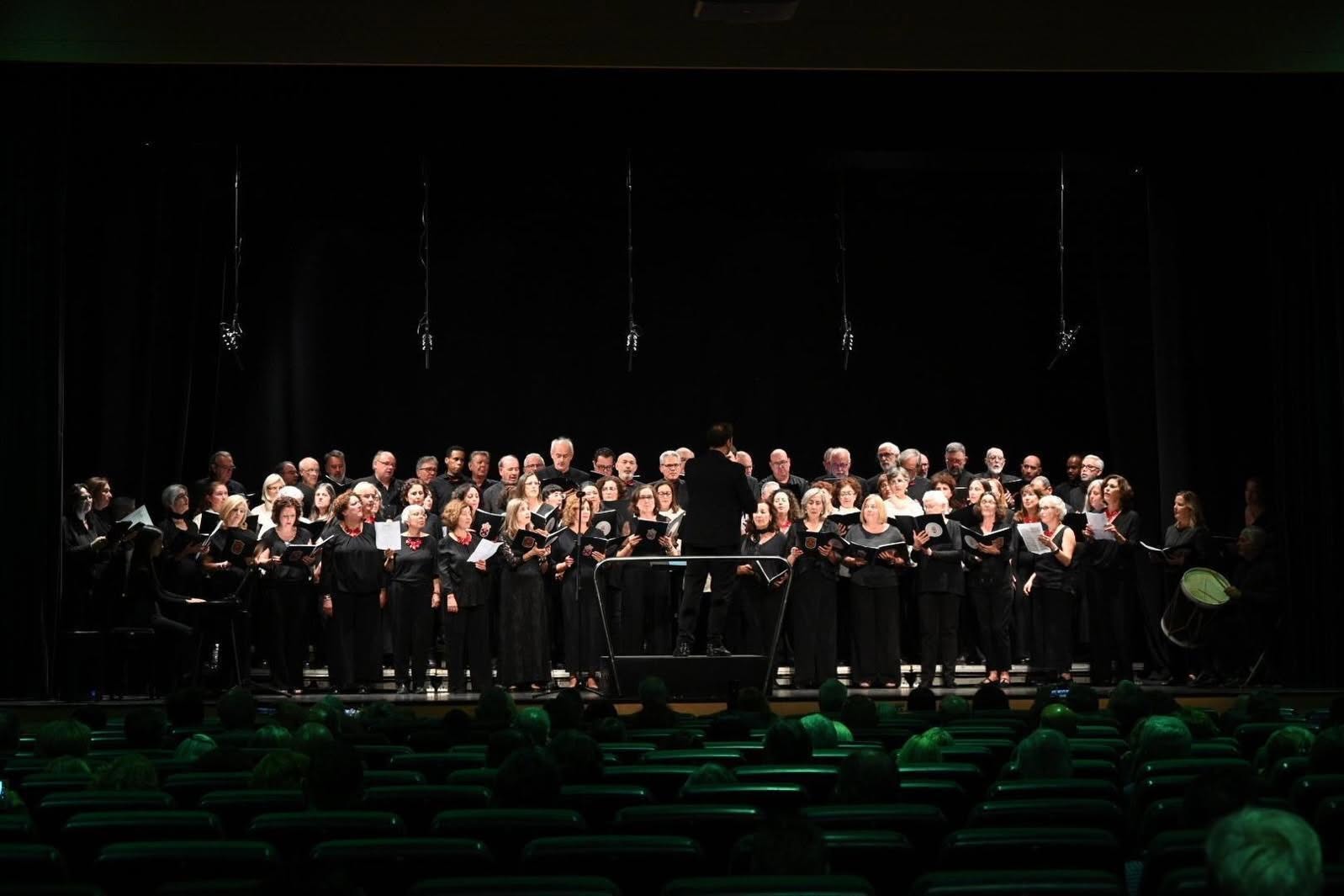 Un coro de personas vestidas de negro, de pie en un escenario con partituras, ante butacas verdes.