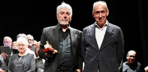 Dos hombres trajeados posan en un escenario; uno sostiene una pequeña figura de premio, con público detrás.