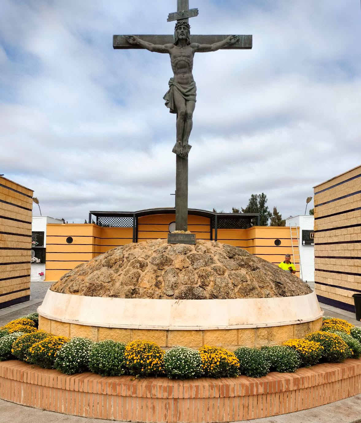 Un gran crucifijo sobre un montículo de piedras, rodeado de flores y edificios amarillos con bandas negras.