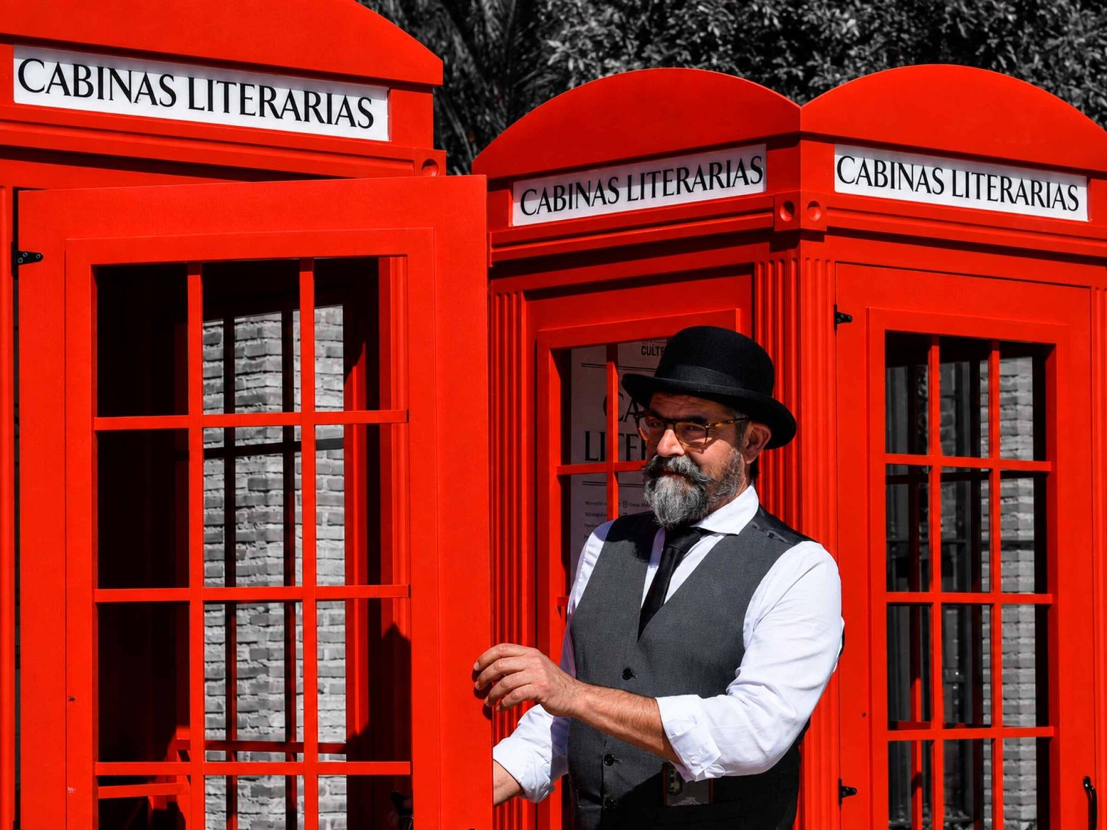 Un hombre con barba, sombrero y chaleco posa junto a tres cabinas rojas rotuladas Cabinas Literarias.