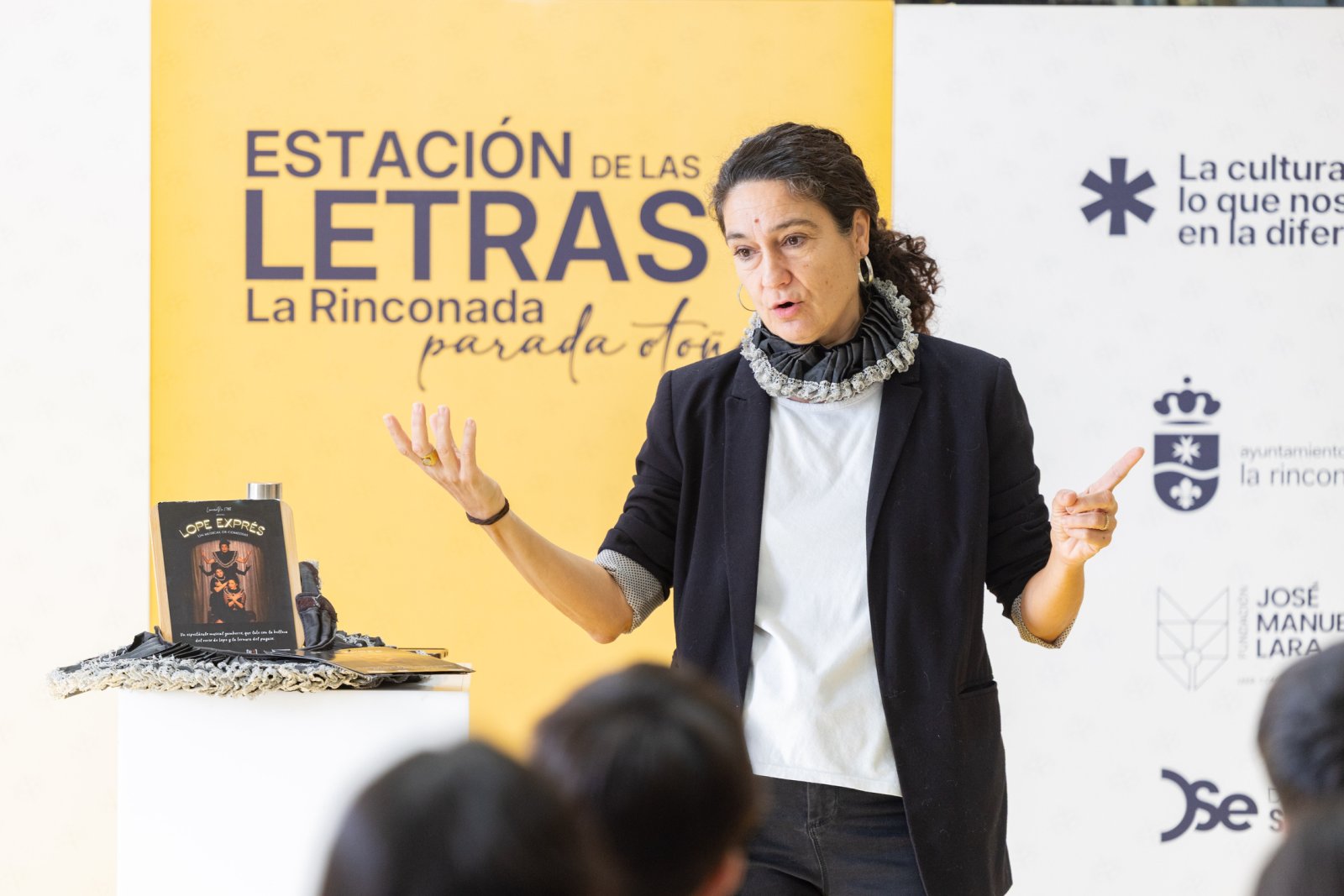 Una mujer con blazer negro y cuello de volantes habla con las manos alzadas ante un público, junto a un atril con un libro y un cartel amarillo de fondo.