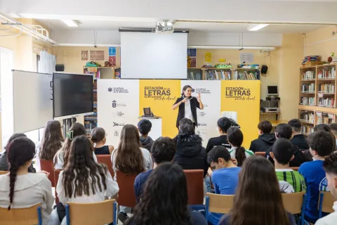 Una mujer habla gesticulando ante un grupo de alumnos sentados de espaldas en un aula con estanterías y un cartel de Estación de las Letras.