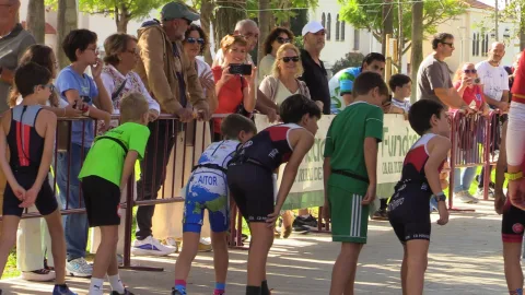 Niños con equipación deportiva esperan junto a una valla metálica ante un grupo de adultos que los observan y hacen fotos en una calle arbolada.