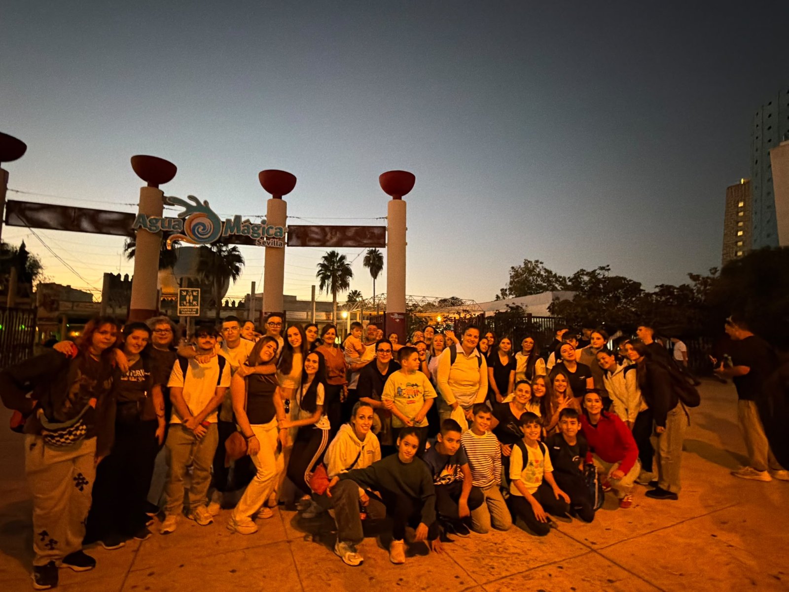 Un grupo numeroso de jóvenes posa sonriente de noche ante la entrada de un parque, bajo un cartel con el nombre del recinto.