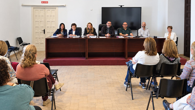 Varias personas sentadas tras una mesa larga frente a un público sentado en sillas, en una sala con pantalla y puerta al fondo.