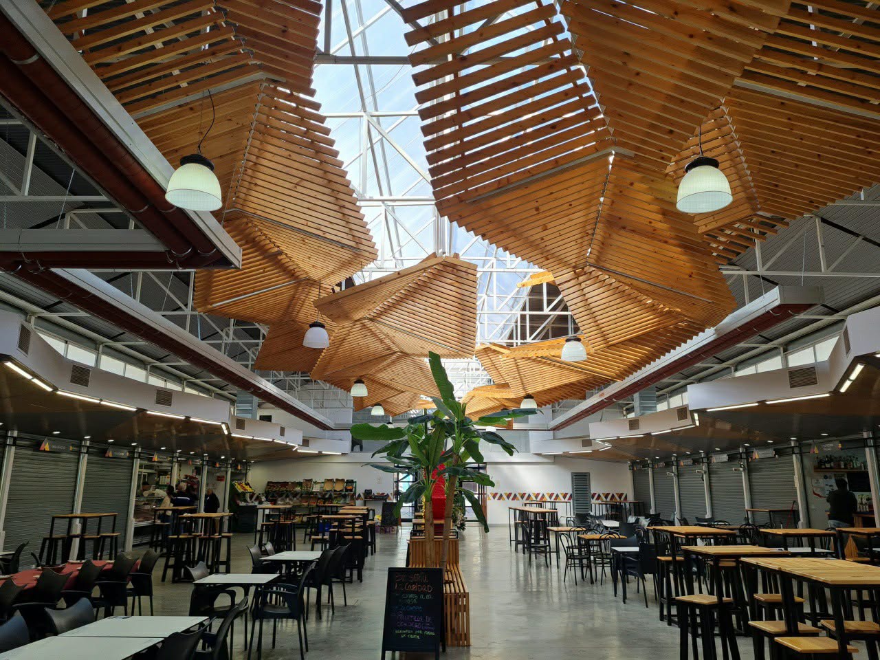 Interior diáfano de un mercado con puestos cerrados, mesas y sillas, y techo de listones de madera bajo una claraboya.