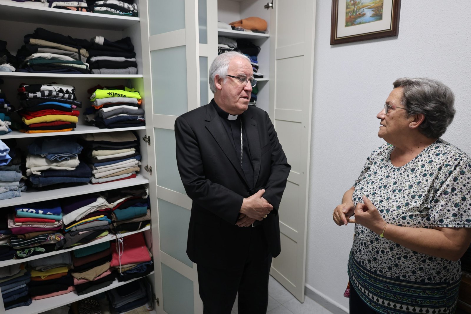 Un hombre con camisa clerical negra y una mujer conversan junto a unas estanterías llenas de ropa doblada.