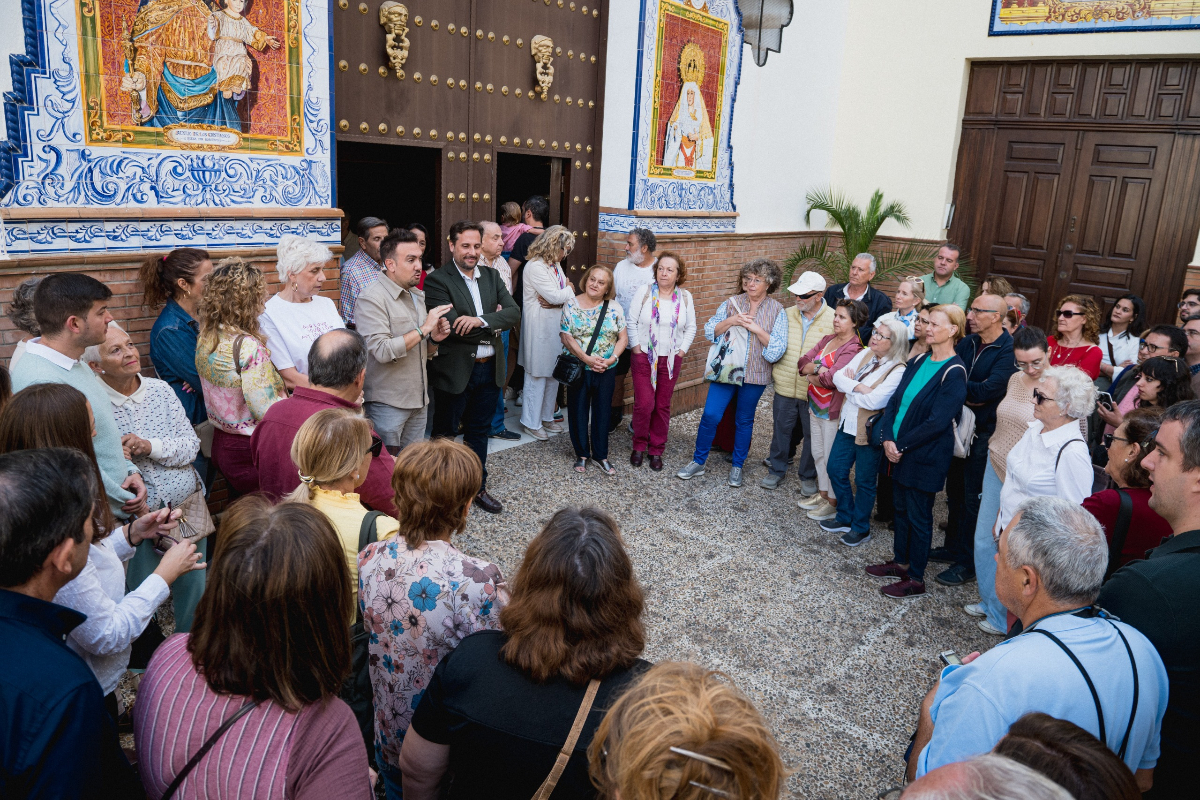 Un grupo de personas rodea en un patio empedrado a un hombre que habla junto a una puerta con azulejos.