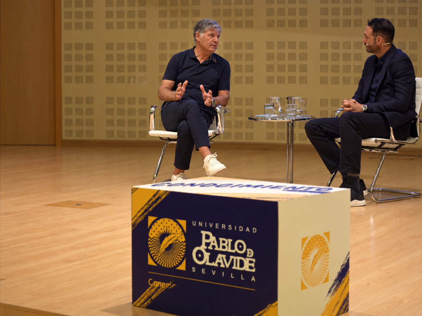 Dos hombres conversan sentados en sillas frente a una mesa con vasos, en un escenario con un cubo de la Universidad Pablo de Olavide.