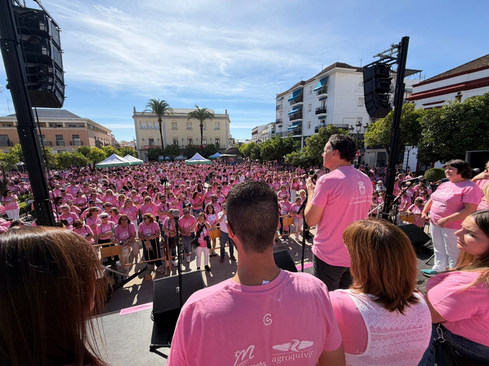 Una multitud con camisetas rosas llena una plaza desde un escenario con altavoces, con edificios y palmeras al fondo.