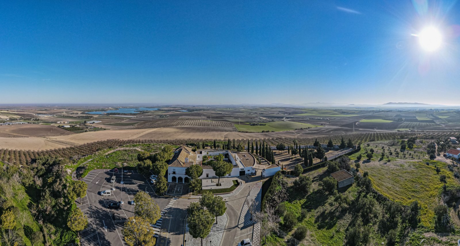 Vista aérea de un edificio blanco con tejados y cipreses junto a un aparcamiento, rodeado de campos de cultivo y un embalse al fondo bajo cielo despejado.