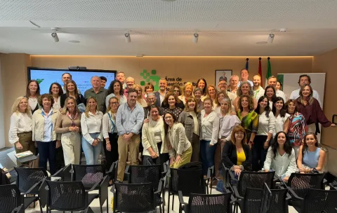 Un grupo numeroso de personas posa de pie y sonriendo en una sala con sillas, pantalla y banderas al fondo.