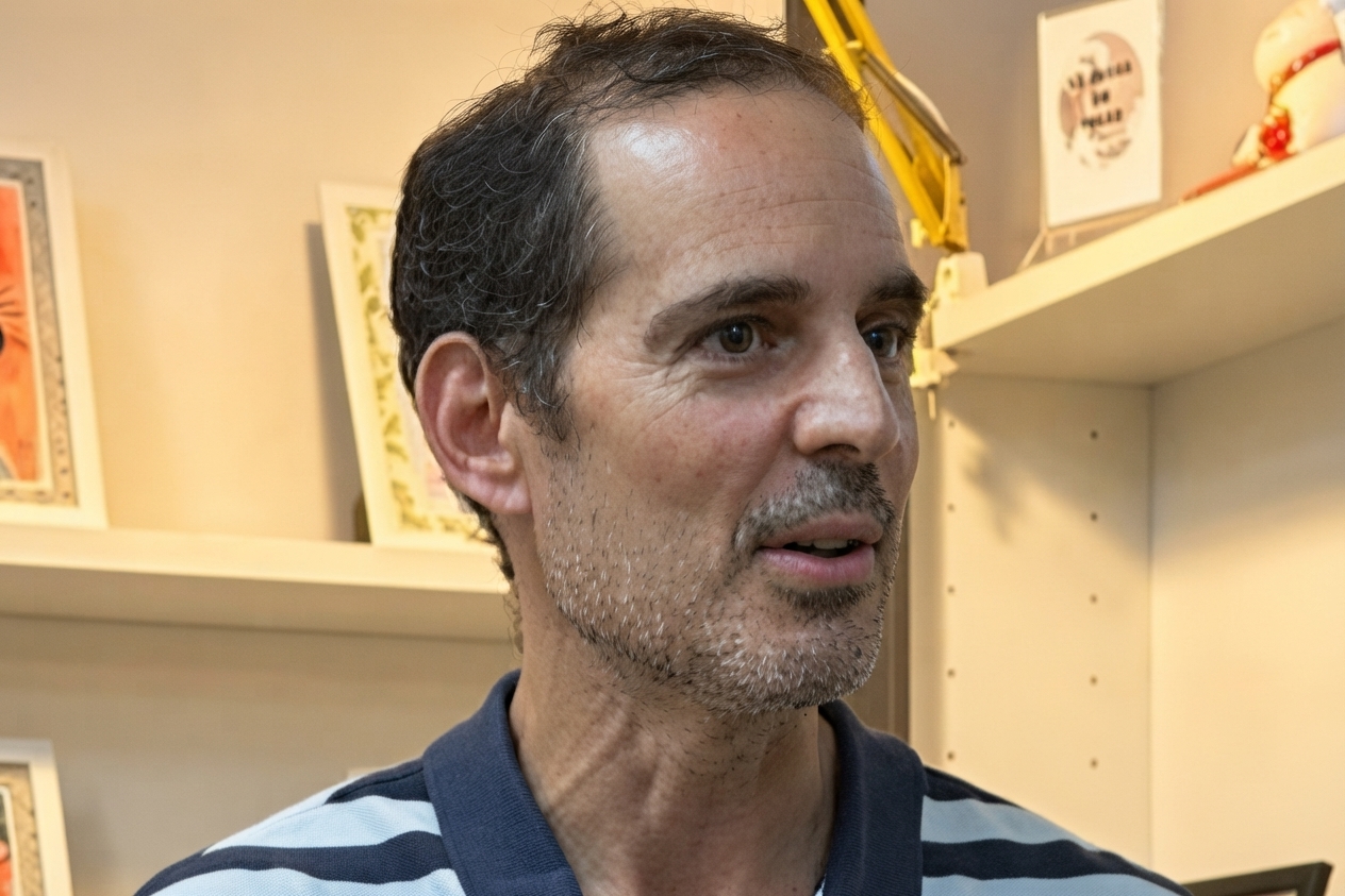 Primer plano de un hombre de mediana edad con barba canosa y polo a rayas, en un interior con estanterías y cuadros.
