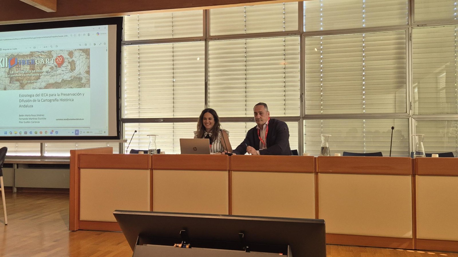 Una mujer y un hombre sentados tras una mesa de madera en una sala con una pantalla que proyecta una presentación sobre cartografía histórica.