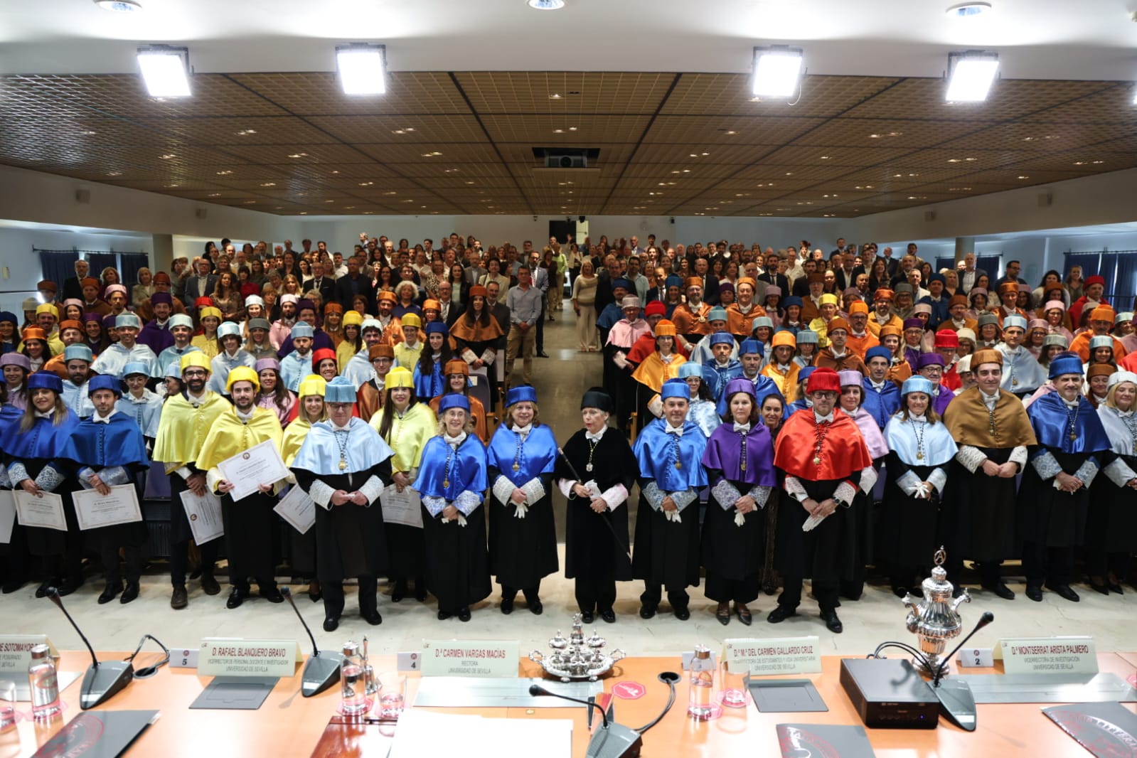 Un numeroso grupo de personas con togas y birretes de colores posa de pie en un salón con mesas y micrófonos al frente.