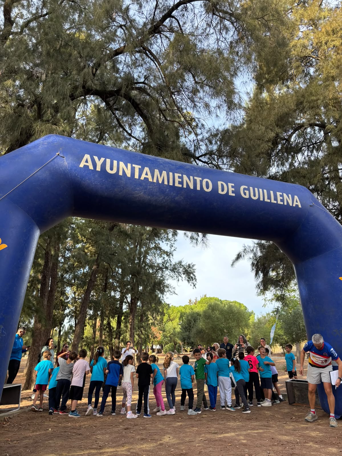 Un grupo de niños y adultos junto a un arco hinchable azul con el texto Ayuntamiento de Guillena, en un entorno arbolado.