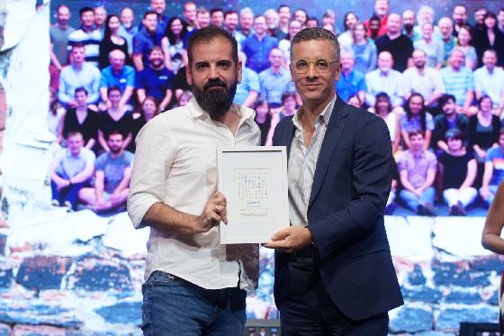 Dos hombres de pie sostienen juntos un diploma enmarcado ante un fondo con una multitud.