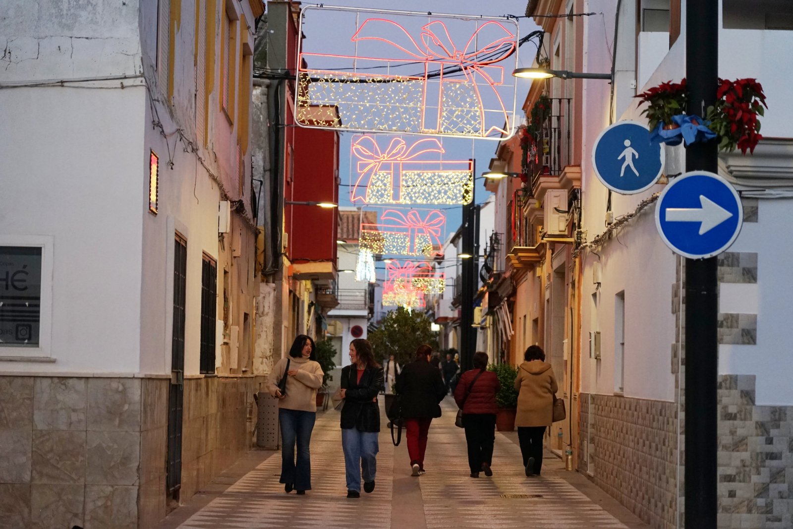 Una calle estrecha con varias personas caminando y luces navideñas con forma de regalos colgadas sobre ella.