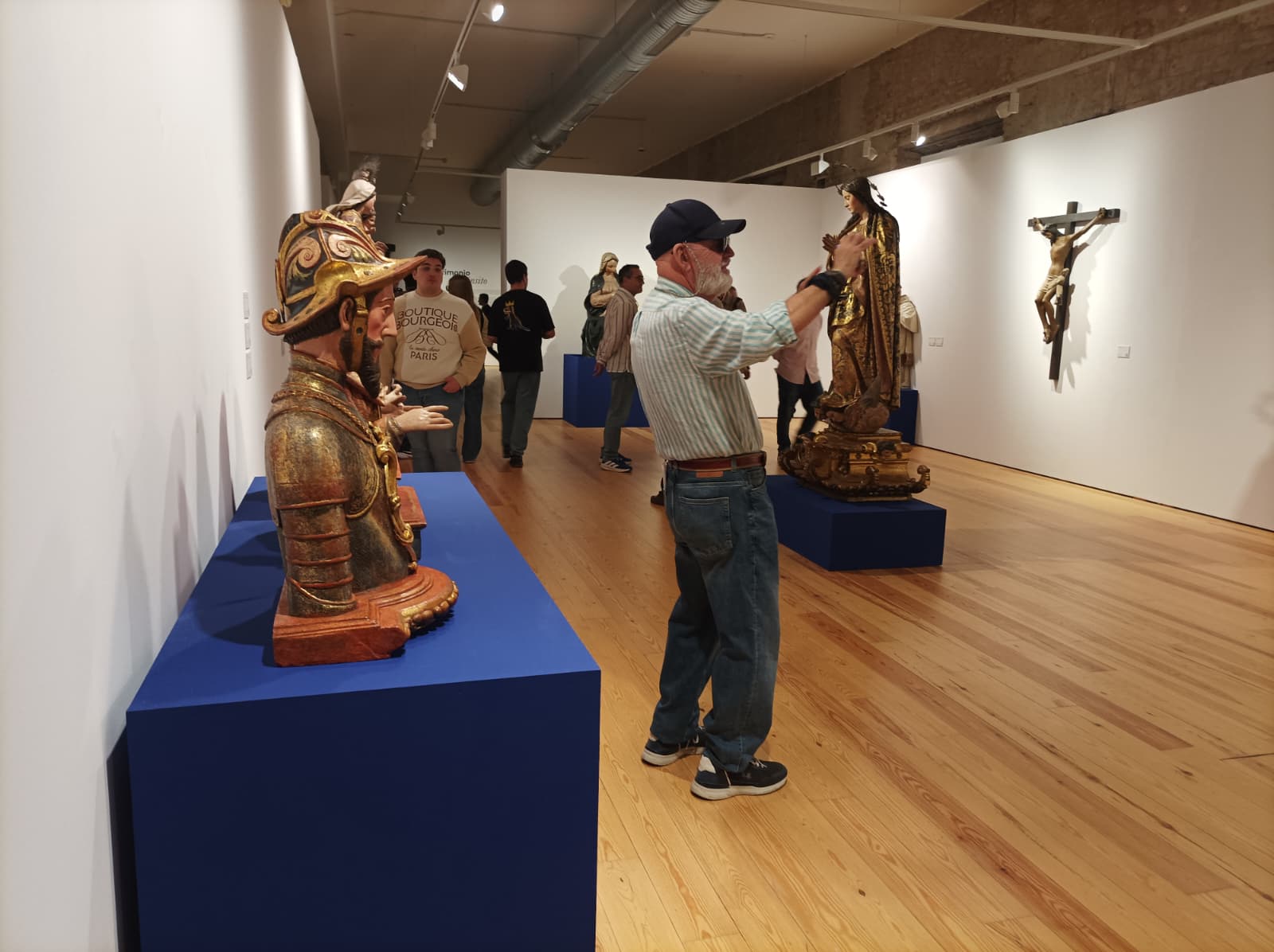 Un hombre con gorra y camisa de rayas fotografía con el móvil una talla dorada en una sala de exposiciones con varias esculturas religiosas sobre peanas azules.