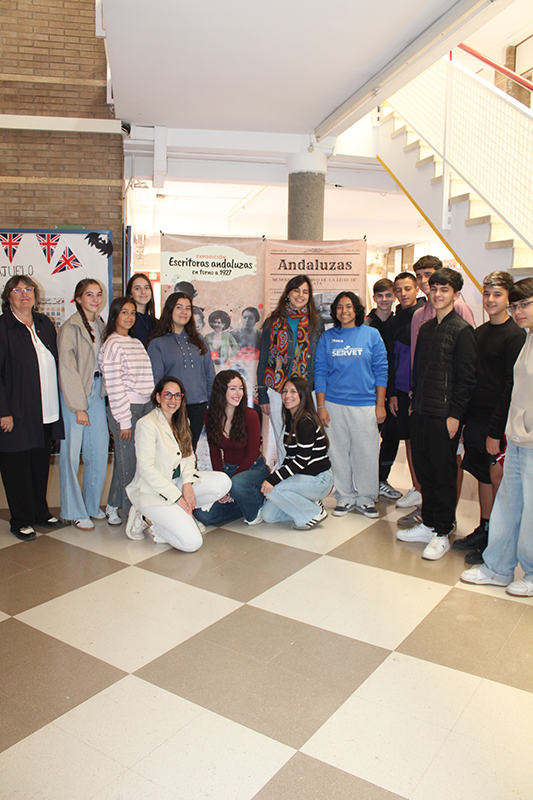 Un grupo de jóvenes y una mujer posan sonrientes junto a carteles de una exposición en un vestíbulo con escaleras y suelo ajedrezado.