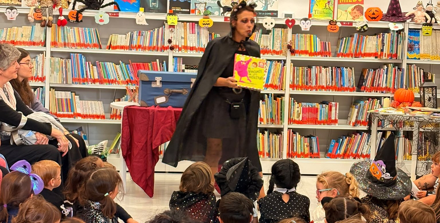 Una mujer disfrazada de bruja muestra un libro a un grupo de niños sentados en una biblioteca decorada con motivos de Halloween.