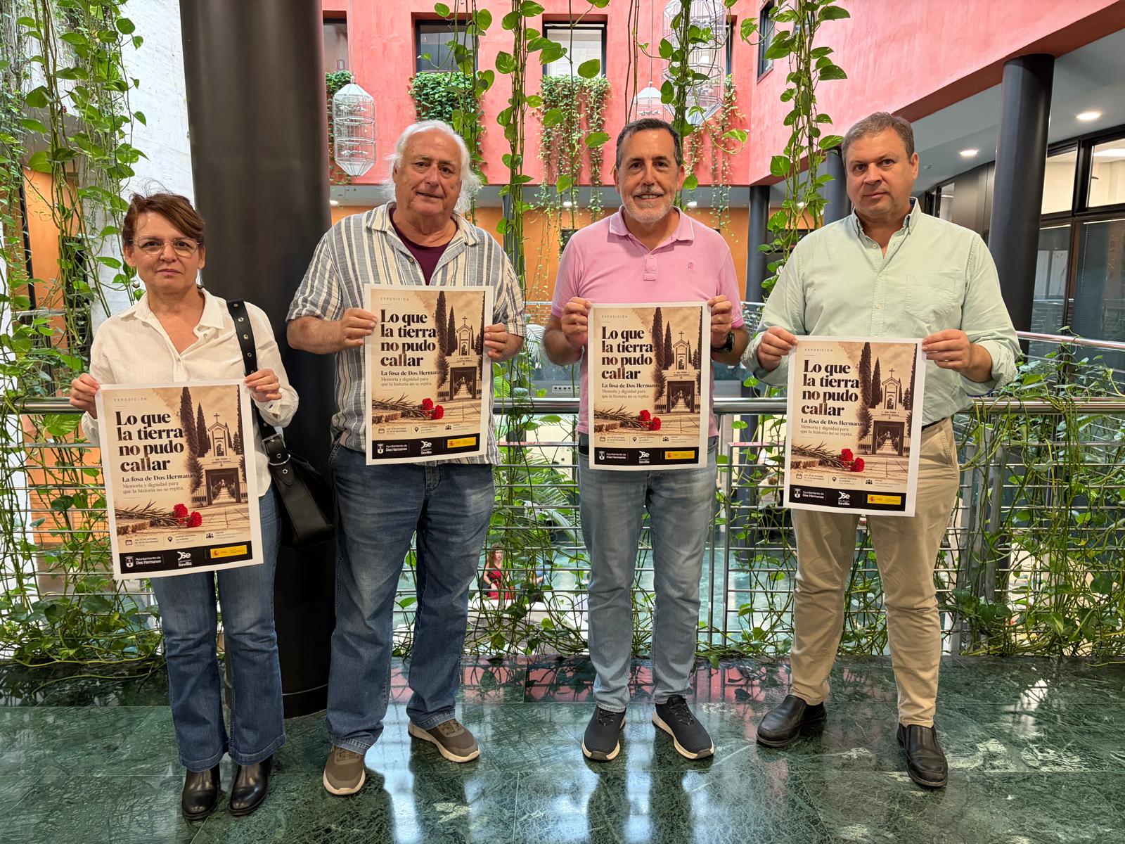 Cuatro personas de pie sostienen carteles de una exposición en un patio interior con plantas y barandilla metálica.