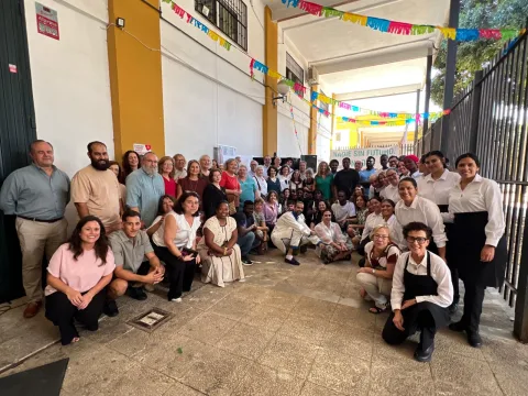 Un grupo numeroso de personas de distintas edades posa sonriente en un patio con banderines de colores y una pancarta al fondo.