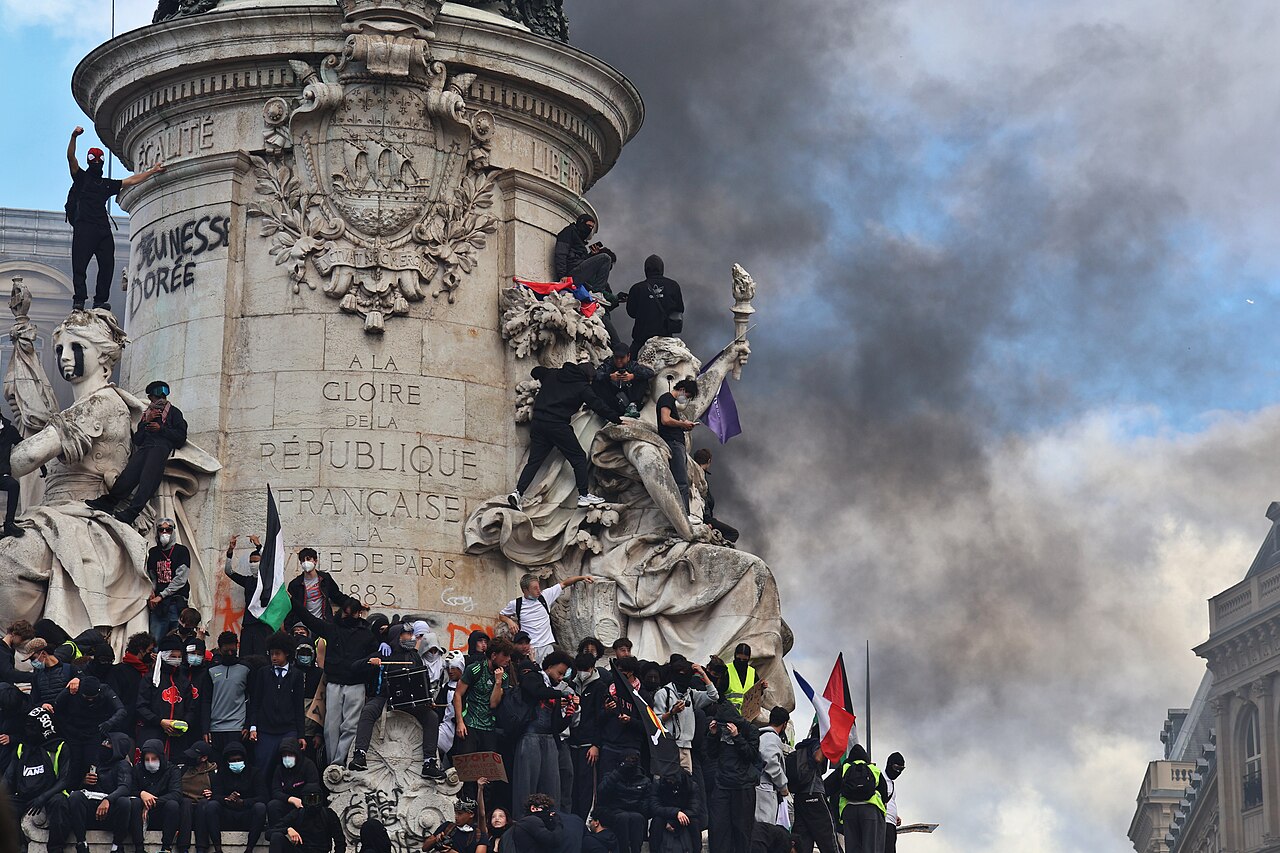 Multitud de personas subidas a un monumento de piedra con inscripciones, ante una gran columna de humo negro.