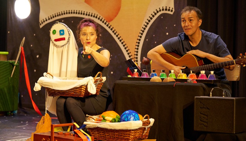 Dos artistas en un escenario: una mujer con un muñeco de trapo y un hombre tocando la guitarra, con cestas y objetos de colores.