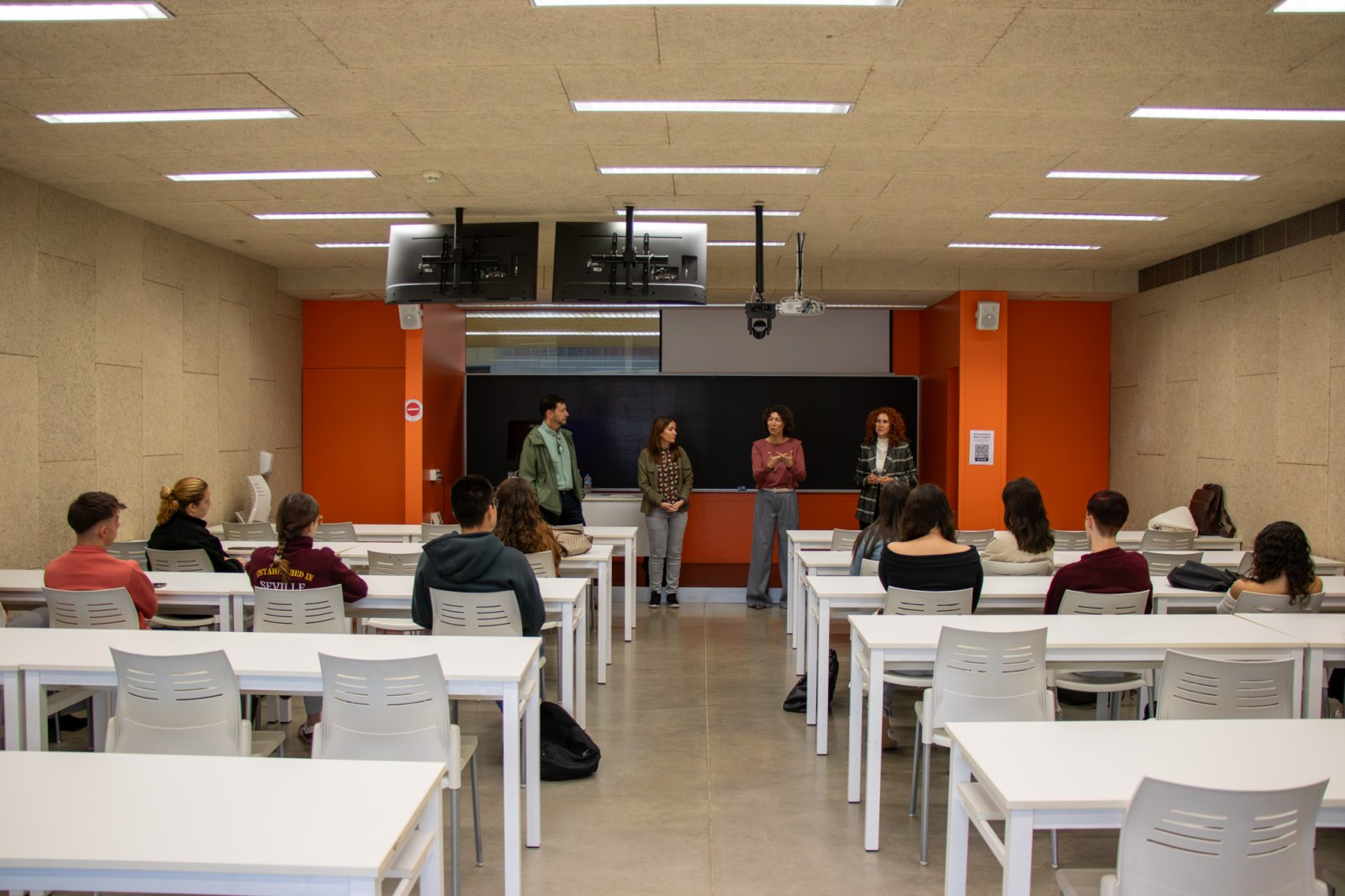 Cuatro personas de pie ante una pizarra en un aula con mesas blancas y alumnos sentados escuchándolas.