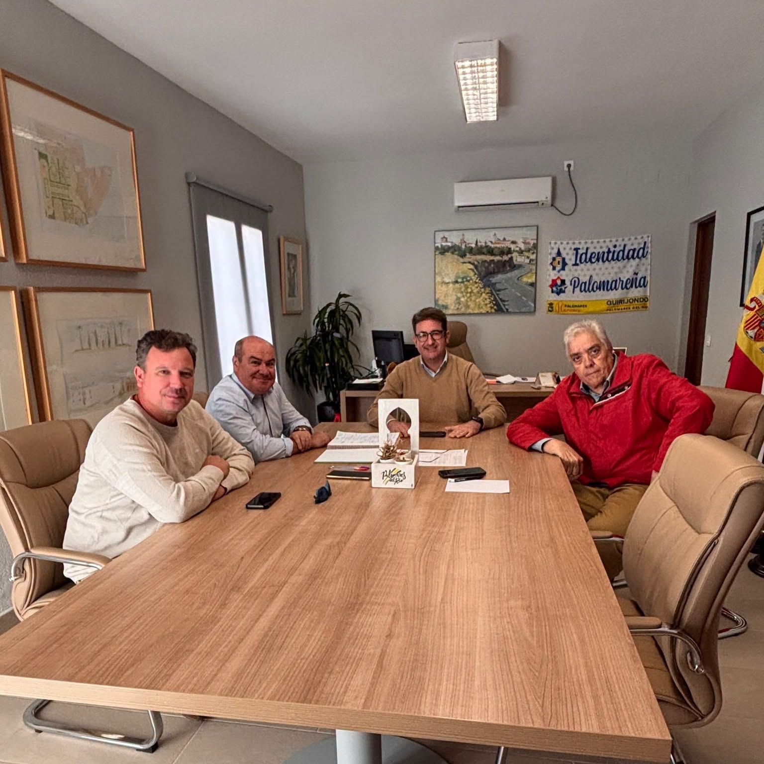 Cuatro hombres sentados en una mesa de reuniones en una sala con carteles y un banderín en la pared.