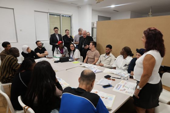 Un grupo de personas sentadas alrededor de una mesa grande en una sala, con varias de pie al fondo y una mujer de pie a la derecha.